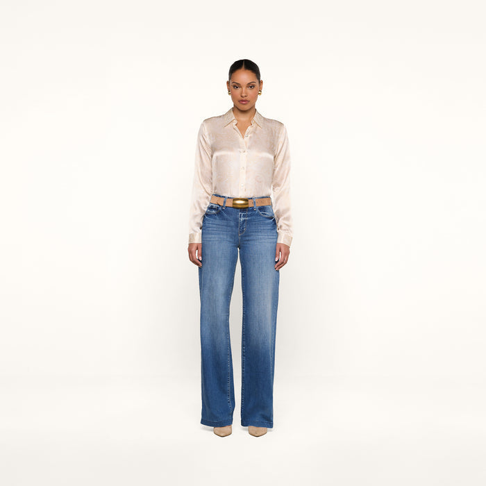 A woman stands facing forward, wearing the Tyler Silk Blouse from L’AGENCE tucked into high-waisted blue jeans and beige heels, against a plain white background.