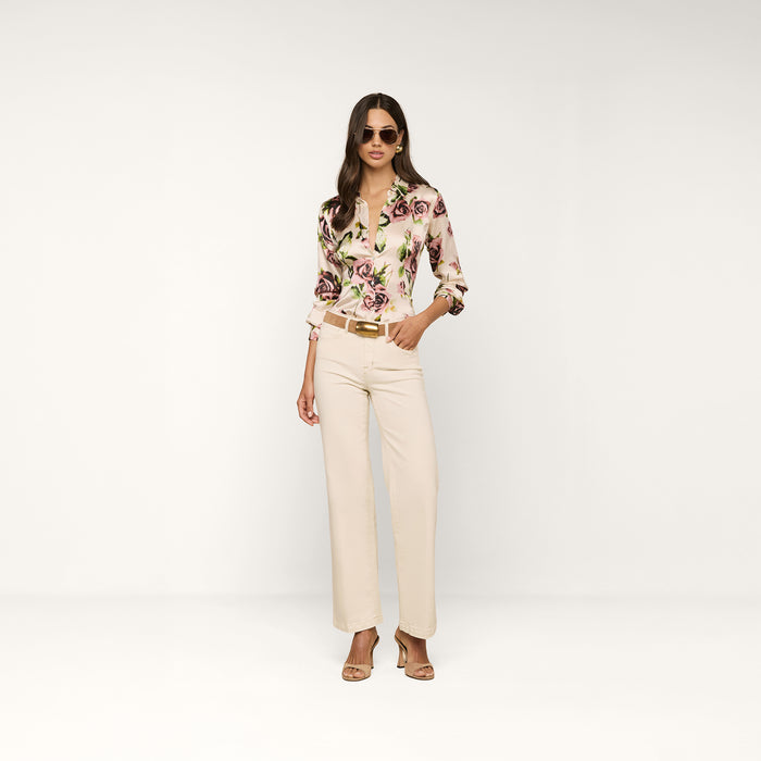 A woman wearing sunglasses, the Tyler Silk Blouse featuring a mauve rose print and long sleeves, beige high-waisted trousers, and heeled sandals poses confidently against a plain white background.