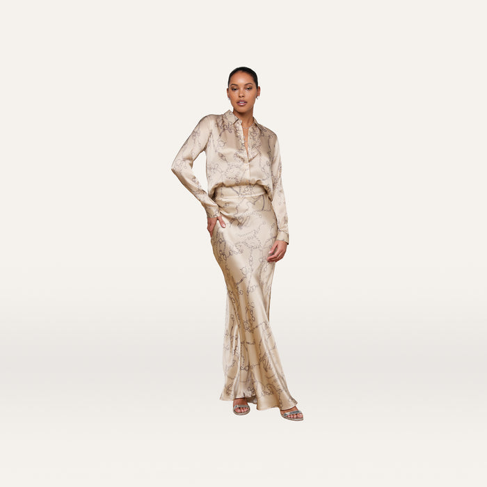 A woman stands against a plain white background, wearing the L’AGENCE Tyler Silk Blouse in a light beige chain print with a matching floor-length skirt and open-toe sandals. She has one hand on her hip and looks confidently at the camera.