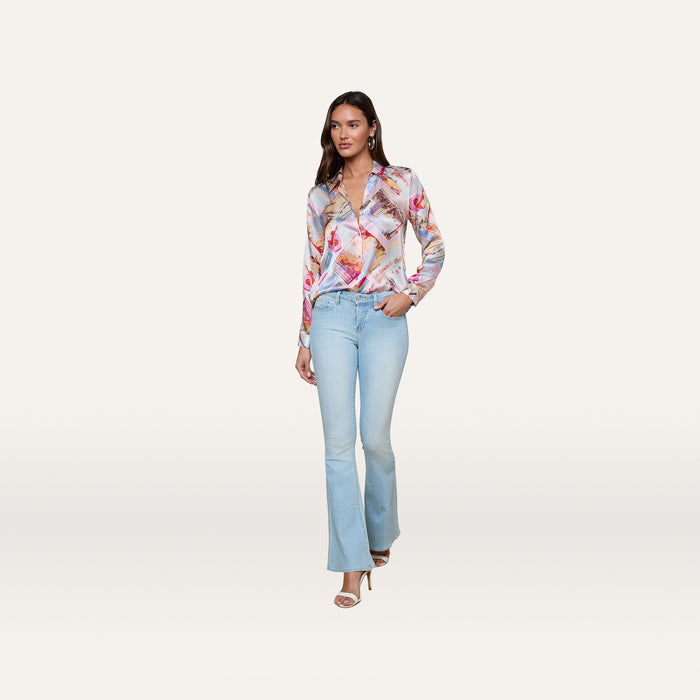 A woman stands confidently against a plain white background, wearing the colorful Tyler Silk Blouse from L’AGENCE paired with light blue flared jeans, her hand in her pocket and open-toed heels.