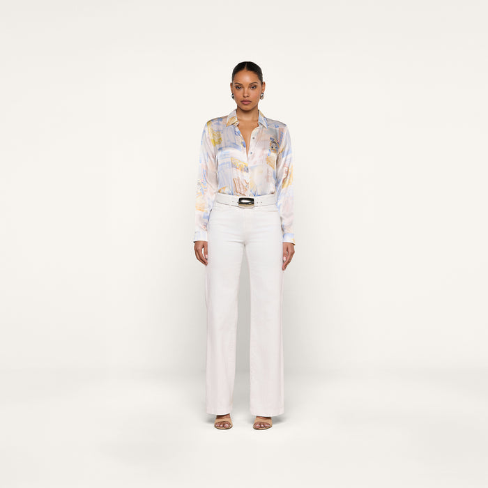 A woman stands facing forward against a plain white background, wearing the Tyler Silk Blouse—a pastel patterned silk button-down—tucked into high-waisted white pants, styled with sandals and a black belt.