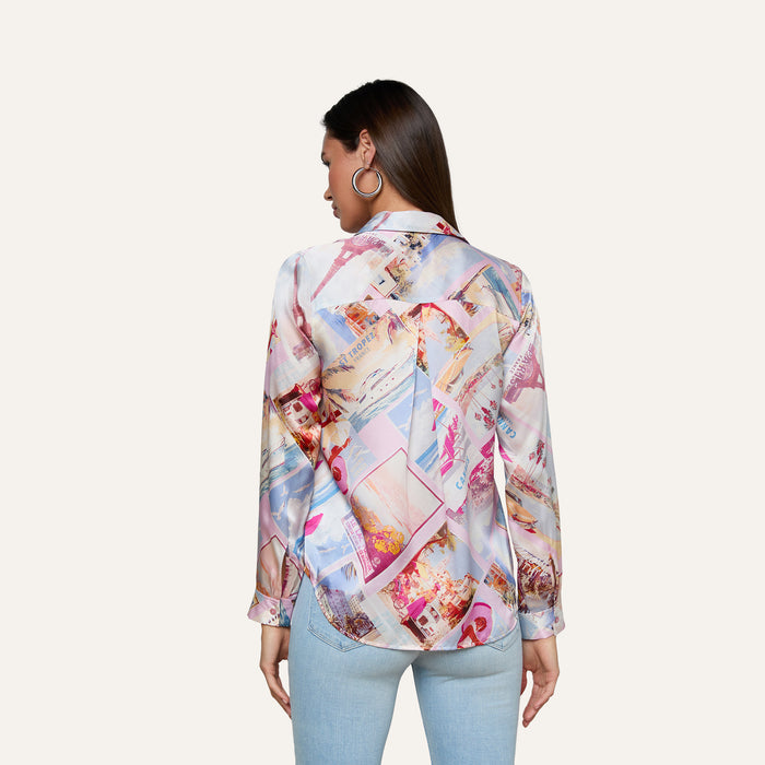 A woman faces away, wearing the Tyler Silk Blouse by L’AGENCE featuring a pink postcard print, paired with light blue jeans. Her dark hair is styled down, and she accessorizes with large hoop earrings. The background is plain and light.