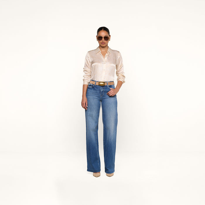 A person wearing sunglasses, the Tyler Silk Blouse in a paisley print, blue wide-leg jeans, and beige heels stands against a plain white background.