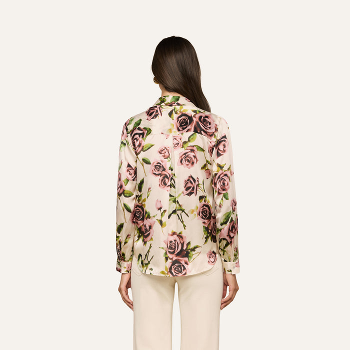 A woman with long brown hair is shown from the back against a plain light background, wearing the Tyler Silk Blouse—a cream button-down blouse featuring a mauve rose print—paired with cream trousers.