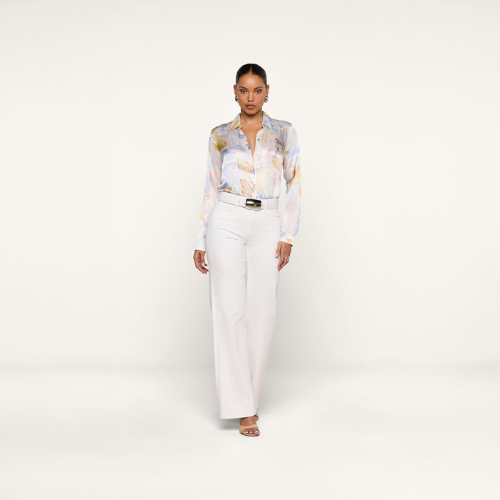 A woman stands facing forward, wearing the L’AGENCE Tyler Silk Blouse with a pastel pattern, tucked into high-waisted white wide-leg pants styled with a black belt and heeled sandals, against a plain white background.