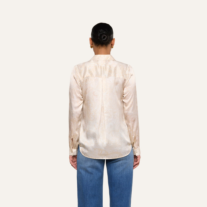 A person stands facing away from the camera, wearing the Tyler Silk Blouse—a cream-colored satin top with a subtle paisley print—paired with blue jeans, against a plain light background.