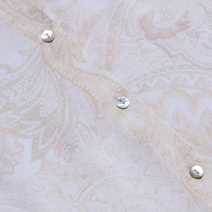 Close-up of the pale, paisley-patterned silk fabric of the Tyler Silk Blouse, featuring three iridescent buttons arranged diagonally. The soft beige and cream tones are adorned with subtle floral and leaf motifs, capturing L’AGENCE’s signature elegance.