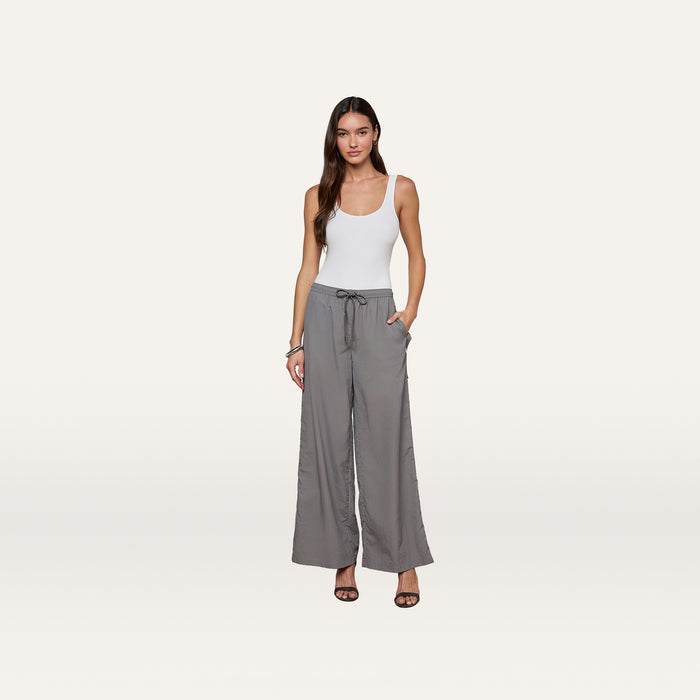A woman stands against a white background, wearing a white tank top, the Valkyrie Snap Pant in gray featuring a wide-leg silhouette and drawstring waist, paired with black heeled sandals and her hands in her pockets.