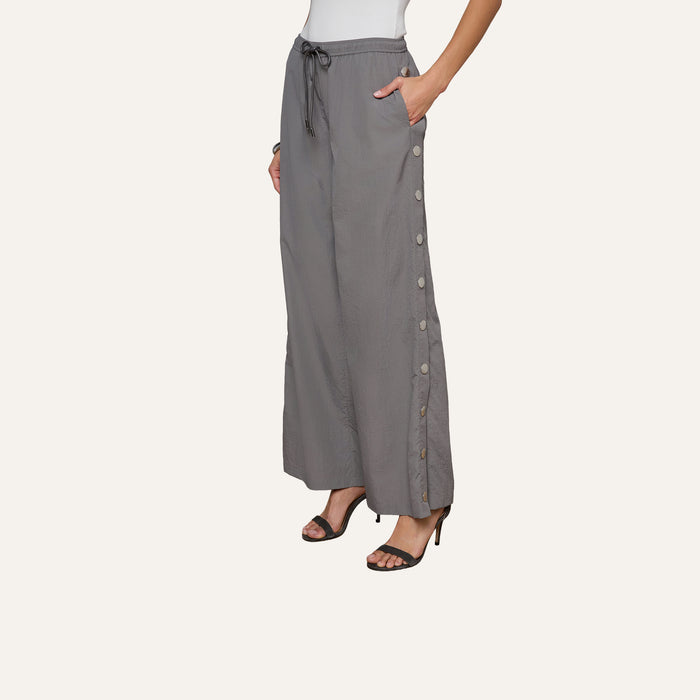 A woman wears loose grey Valkyrie Snap Pants with side buttons, paired with black high-heeled sandals and a white sleeveless top. Her left hand rests in her pocket, and the background is plain off-white.