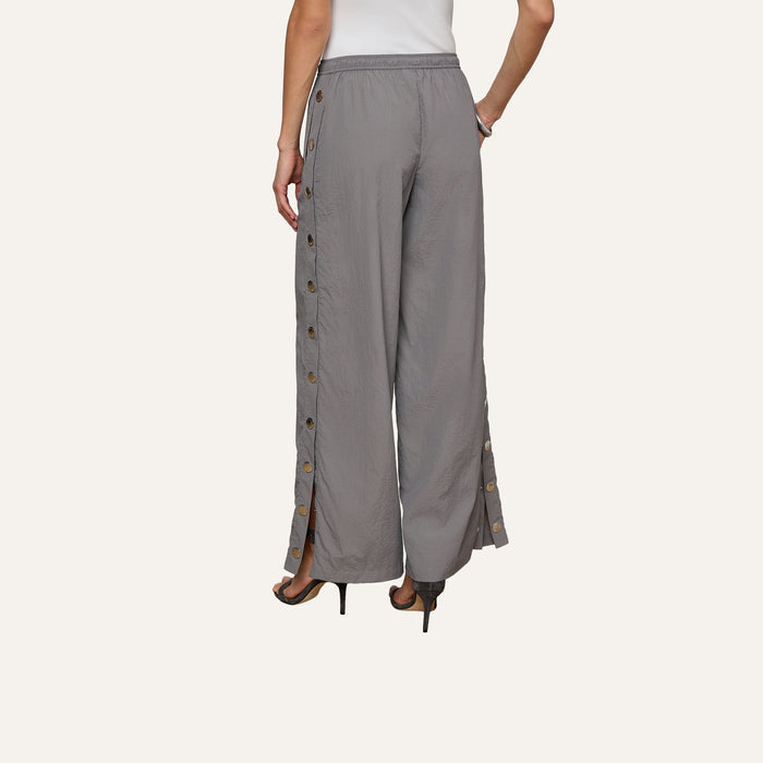 Person wearing loose gray Valkyrie Snap Pant with side buttons and black heels, standing with one hand on hip. Back view against a plain background.