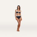 A woman stands barefoot wearing the Vanessa High-Waisted Bikini Bottom in navy blue with white trim and gold button details, crafted from textured bouclé fabric. She faces forward with her arms relaxed by her sides, set against a plain light background.