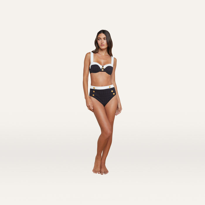 A woman stands barefoot wearing the Vanessa High-Waisted Bikini Bottom in navy blue with white trim and gold button details, crafted from textured bouclé fabric. She faces forward with her arms relaxed by her sides, set against a plain light background.