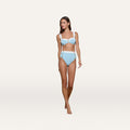 A woman stands wearing the Vanessa High-Waisted Bikini Bottom, a retro-inspired light blue piece crafted from bouclé swim fabric with white straps and accents for moderate coverage, set against a plain white background.