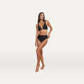 A woman stands on one leg against a plain white background, wearing the Vanessa High-Waisted Bikini Bottom with moderate coverage and a V-neck bikini top.