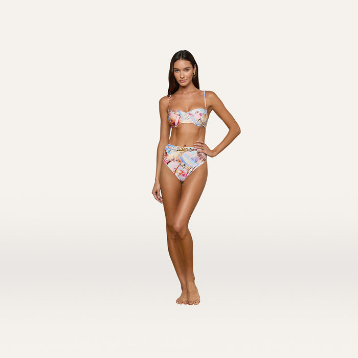 A woman stands barefoot, wearing the Vanessa High-Waisted Bikini Bottom as part of a colorful, postcard print bikini set with a cropped top, posing against a plain, light background.