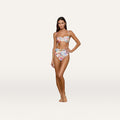 A woman stands barefoot, wearing the Vanessa High-Waisted Bikini Bottom as part of a colorful, postcard print bikini set with a cropped top, posing against a plain, light background.