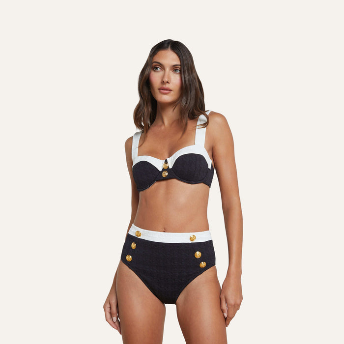 A woman wears the Vanessa High-Waisted Bikini Bottom in navy blue and white with gold buttons. The structured top is crafted from textured bouclé fabric, and she stands against a plain, light background.
