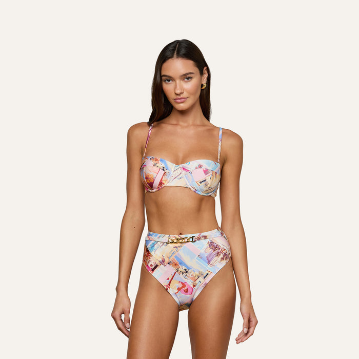 A woman wears the Vanessa High-Waisted Bikini Bottom, part of a colorful postcard print bikini with high-rise bottoms and thin straps, standing against a plain light background and facing forward with a neutral expression.