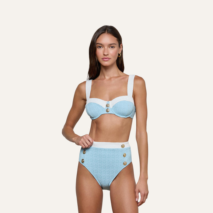 A woman stands against a plain, light background, looking at the camera with a neutral expression while wearing the Vanessa High-Waisted Bikini Bottom in light blue and white bouclé swim fabric, featuring decorative buttons and a supportive fit.