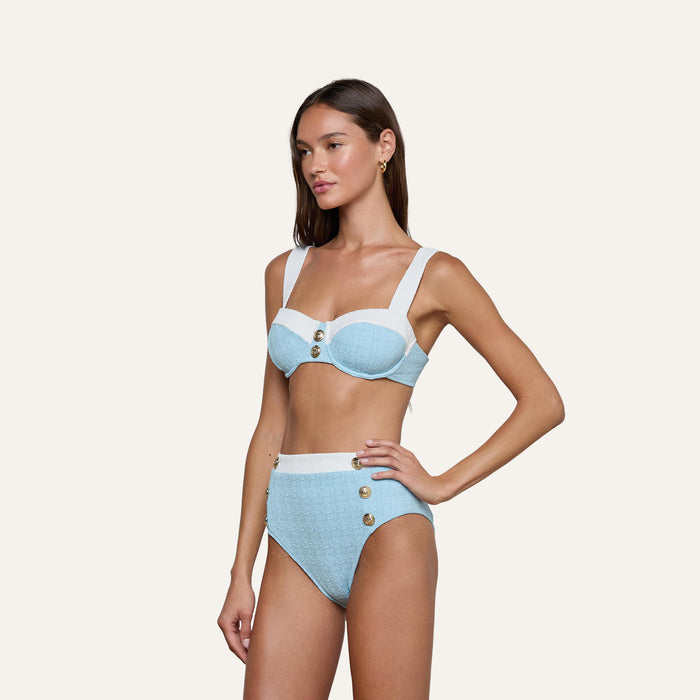 A woman stands against a plain background, wearing the Vanessa High-Waisted Bikini Bottom in light blue and white bouclé swim fabric. The supportive bottoms feature a high-waisted fit with gold button accents, and she poses with one hand on her hip.