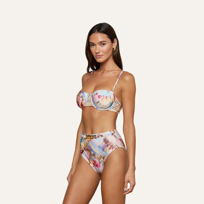 A woman wears the Vanessa High-Waisted Bikini Bottom in a postcard print, paired with thin-strap bikini top, as she stands against an off-white background, facing slightly to the side and looking at the camera.