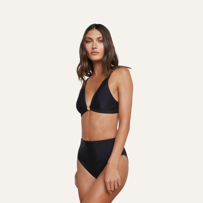 A woman stands against a plain light background wearing the Vanessa High-Waisted Bikini Bottom in black, paired with a plunge top featuring a subtle gold accent, creating a versatile look with moderate coverage.