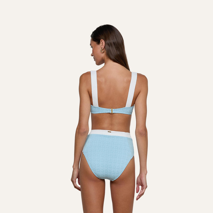 A woman with long brown hair models the Vanessa High-Waisted Bikini Bottom in light blue bouclé swim fabric with white straps and waistband, shown from the back against a plain light background. This supportive bikini bottom offers moderate coverage.