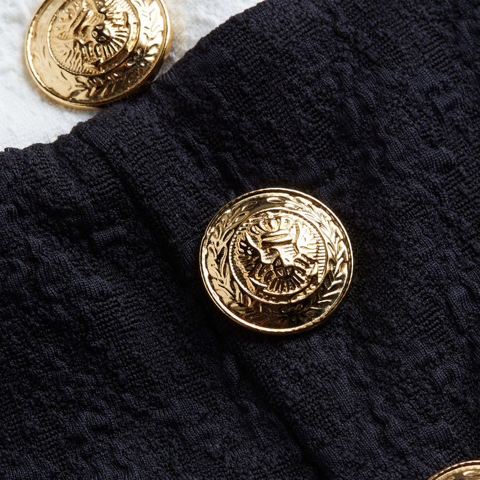 A close-up of a decorative gold button with an eagle design, featured on the Vanessa High-Waisted Bikini Bottom in textured bouclé fabric. Another matching gold button is partially visible on a white surface in the background.