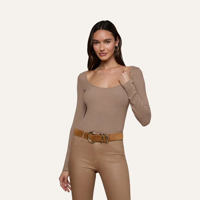 Wearing the Vanya Scoopneck Top in taupe, this woman has it tucked into high-waisted beige pants paired with a wide brown belt, standing against a plain light background and smiling softly.