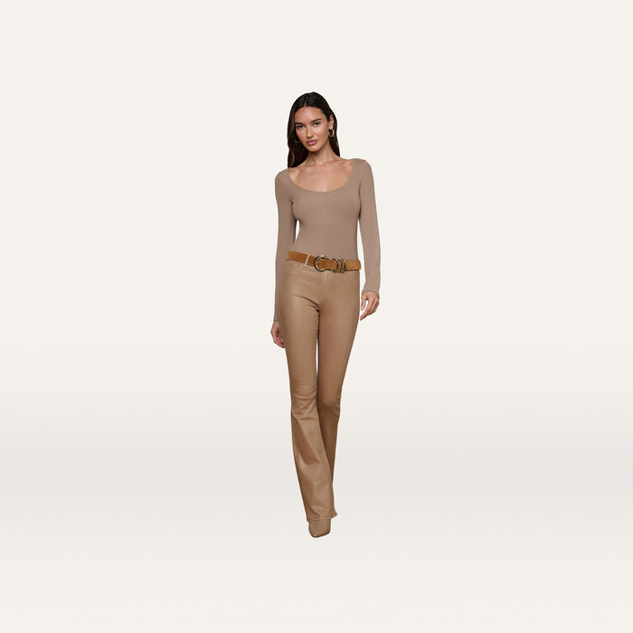 A woman stands against a plain white background, facing forward with a slight smile, wearing the Vanya Scoopneck Top—a fitted tan top crafted from soft stretch rib material—paired with tan pants and a wide brown belt.