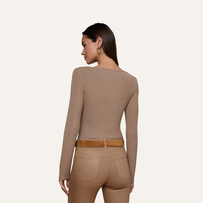 The woman, seen from behind, is wearing the Vanya Scoopneck Top—a fitted, long-sleeve style made from soft stretch rib—tucked into high-waisted tan pants with back pockets and a brown belt. Her straight hair falls loose as she stands against a plain light background.