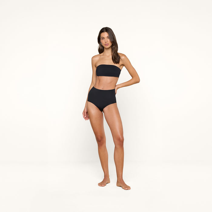 A woman stands barefoot against a plain white background, wearing a black strapless bikini top and the Velle Knit Panty, a high-waisted style that offers the smoothing benefits of compression underwear.