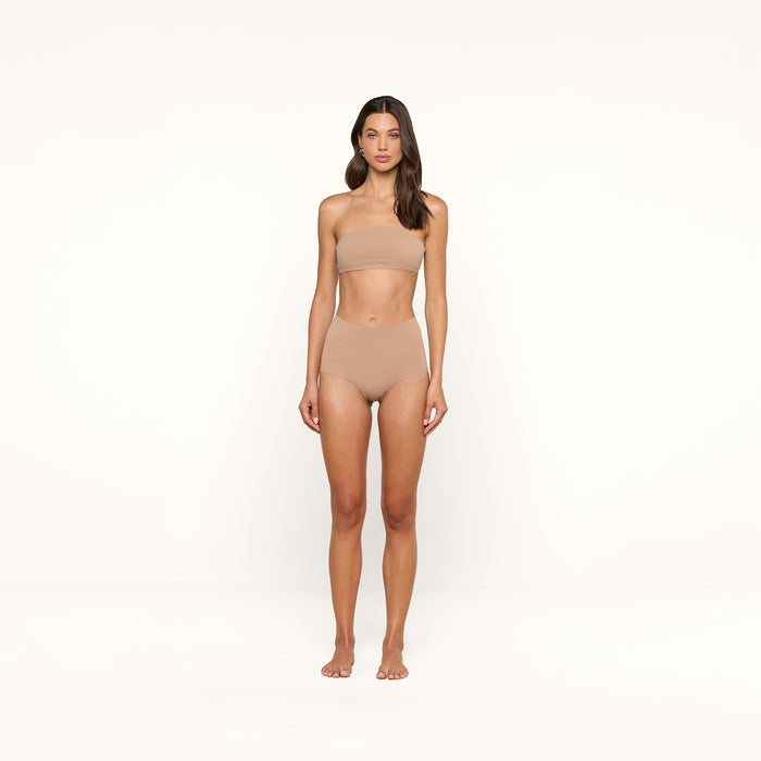 A woman stands facing forward, wearing a nude-colored bandeau top and the Velle Knit Panty, against a plain white background.