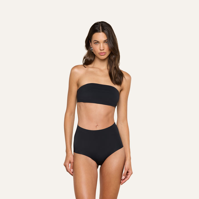 A woman stands and poses in the Velle Knit Panty paired with a black strapless bandeau bikini top against a plain, light background.