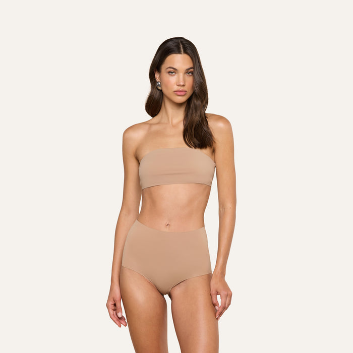 A woman with long, wavy hair and a neutral expression stands against a plain background wearing the Velle Knit Panty paired with a strapless nude bandeau bikini top.