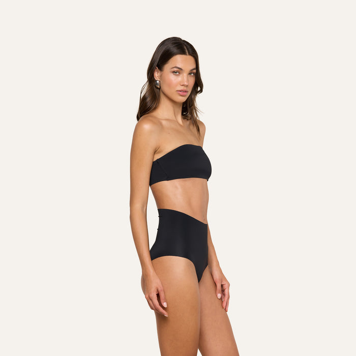 A woman stands and poses against a plain light background wearing the Velle Knit Panty in black, styled as part of a strapless bandeau bikini set with high-waisted bottoms.