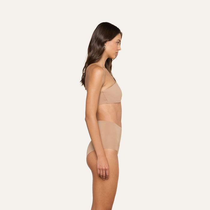 A woman stands in profile against a plain background, wearing a matching nude-colored L’AGENCE Velle Knit Panty and undergarment set.