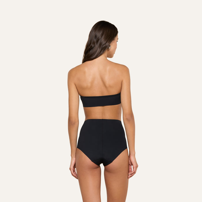 A woman with long brown hair, seen from the back, wears a black strapless bandeau top and the Velle Knit Panty against a plain light background.