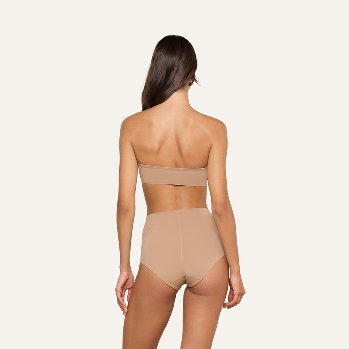 A woman with long brown hair, seen from behind, wears a strapless nude bandeau bra and the Velle Knit Panty against a plain white background.