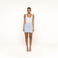 A woman stands facing forward, wearing a white tank top, the Vera Pinstriped Mini Skirt in light blue crafted from a cotton blend, and nude heels, against a plain white background.