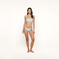 A woman stands on a plain white background, wearing an ice blue Violet String Bikini Bottom with UPF 50 protection. She faces forward with one hand relaxed by her side and the other slightly bent. Her hair is long and loose.