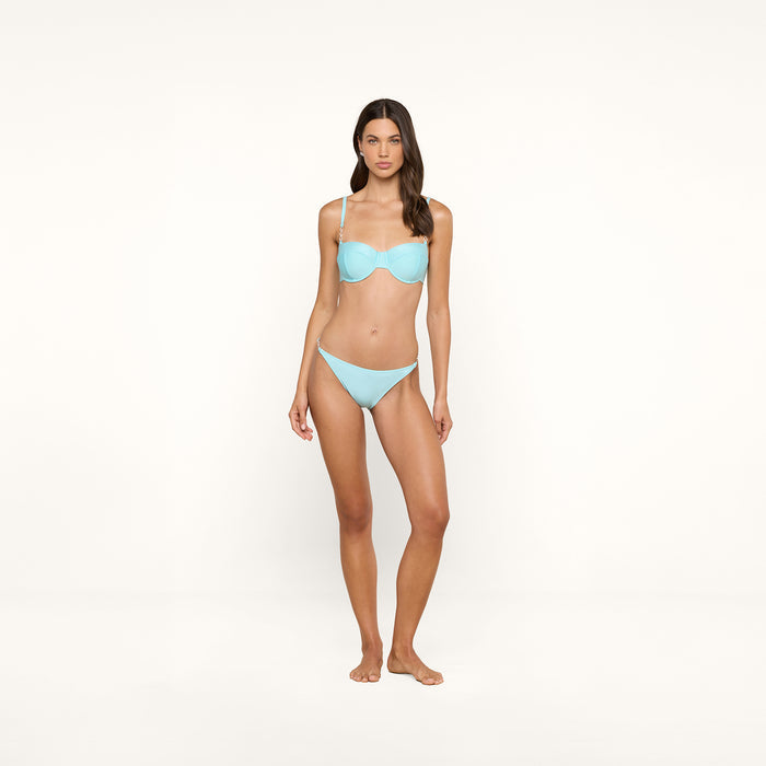 A woman stands on a plain white background, wearing an ice blue Violet String Bikini Bottom with UPF 50 protection. She faces forward with one hand relaxed by her side and the other slightly bent. Her hair is long and loose.
