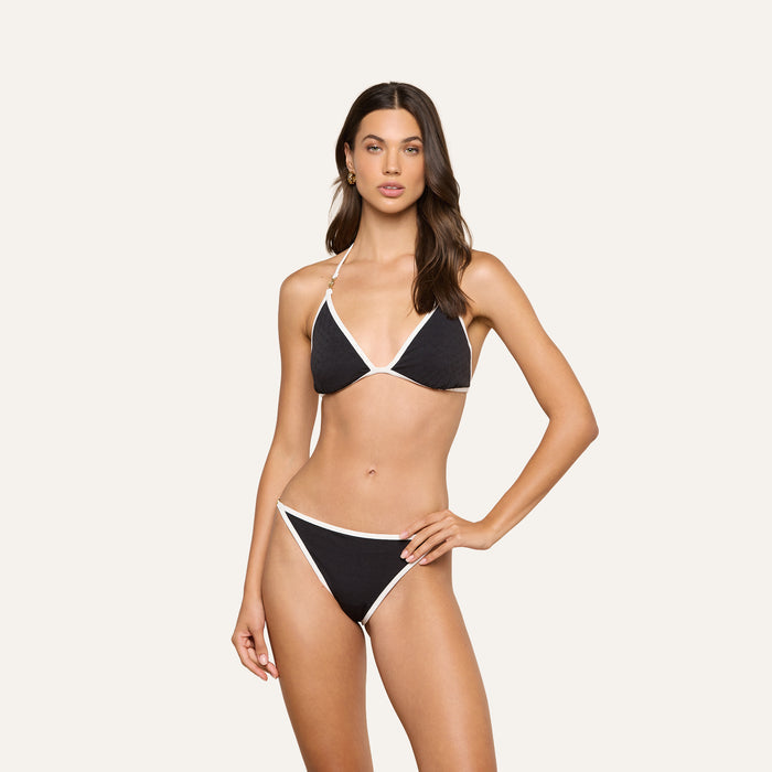 A woman poses wearing a UPF 50 black triangle bikini top with white trim and the Violet String Bikini Bottom against a plain light background.
