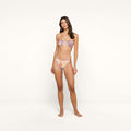 A woman stands barefoot against a plain white background, wearing a colorful palm leaf print bikini featuring the Violet String Bikini Bottom. She has long brown hair and a neutral expression, with her arms relaxed by her sides. The bikini offers UPF 50 sun protection.