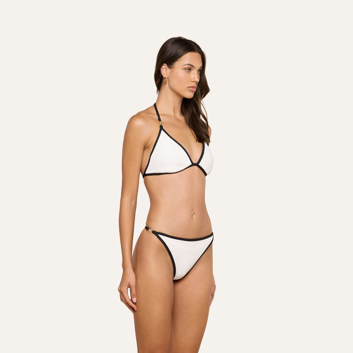 A woman stands against a plain, light background wearing the Violet String Bikini Bottom and a white bikini top with black trim. She is looking slightly to the side with a neutral expression.