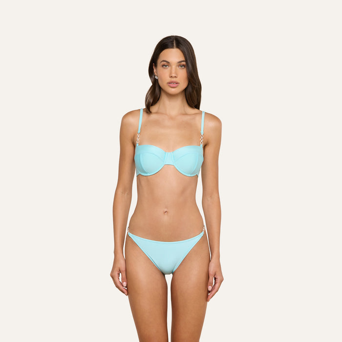 A woman stands facing forward, wearing the Violet String Bikini Bottom in ice blue with gold chain details at the hips, against a plain, light background.