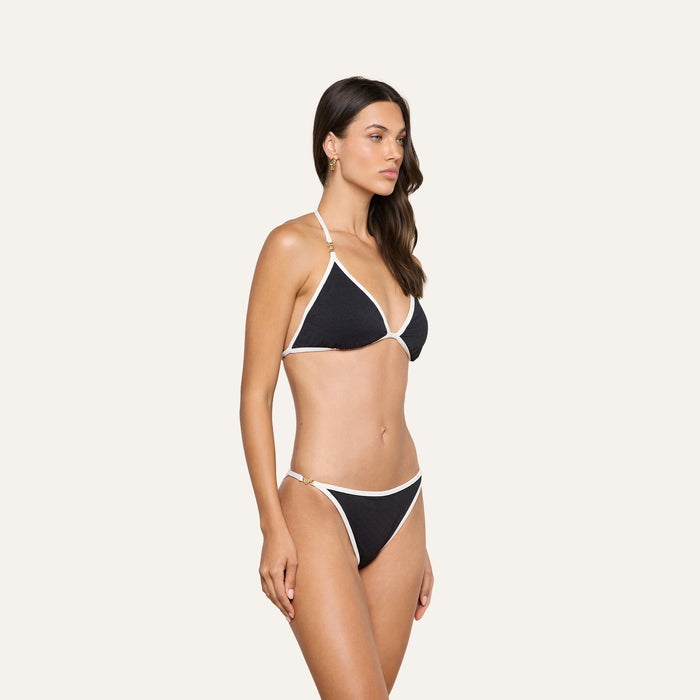 A woman stands against a plain light background, facing slightly to the right with her arms at her sides, wearing the Violet String Bikini Bottom and a black bikini top with white trim.