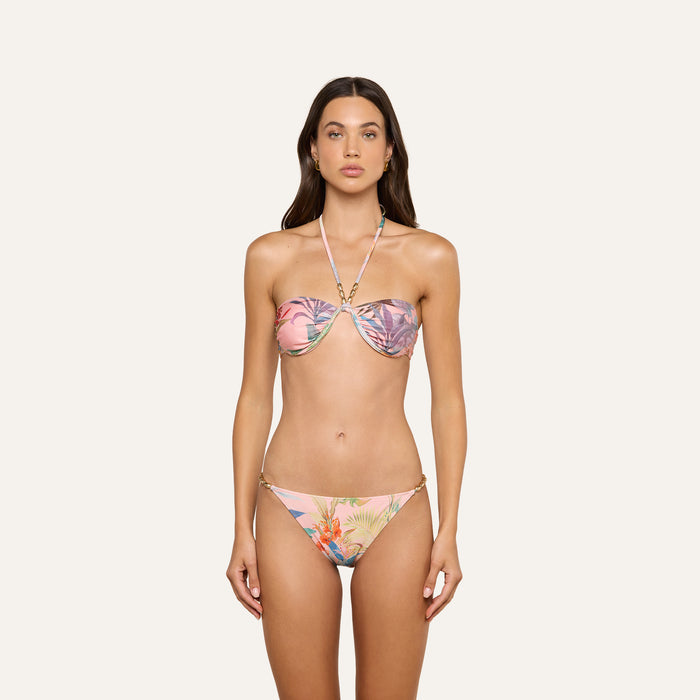 A woman is standing facing forward, wearing the Violet String Bikini Bottom and a pink floral strapless bikini top with a halter neck strap. The background is plain and light-colored.