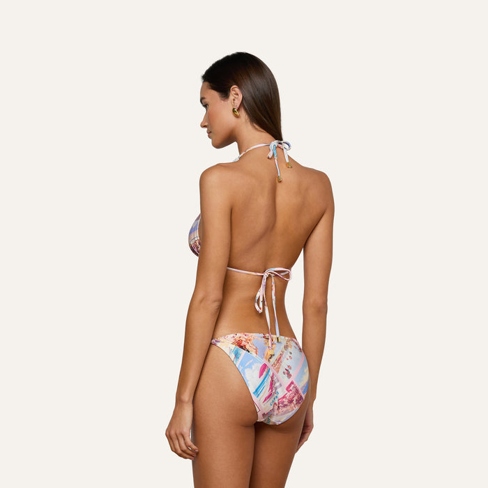 A woman stands with her back to the camera wearing the Violet String Bikini Bottom in a vibrant postcard print, paired with a colorful halter bikini top, set against a plain, light background.