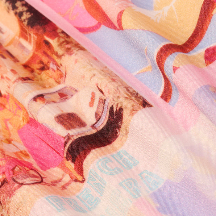 A close-up of the Violet String Bikini Bottom reveals its colorful fabric adorned with abstract illustrations—including a face, wavy shapes, and partially visible light blue text that reads “FRENCH RIVIERA.” The vibrant design channels the playful spirit of a postcard print.